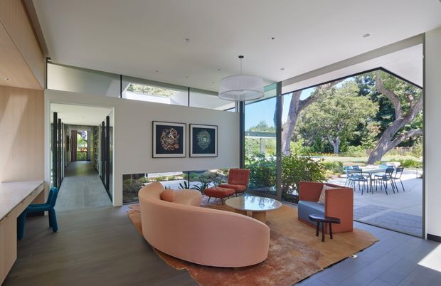 A bright contemporary living room with floor-to-ceiling glazing opening onto a paved outdoor terrace and landscaped garden. The room features a curved blush-pink sofa, rust and blue lounge chairs, a round glass coffee table, a large area rug, timber wall panelling and built-in cabinetry, framed artwork, and a sculptural pendant light. A long hallway extends from the living area, while an outdoor dining setting is visible through the glazing.