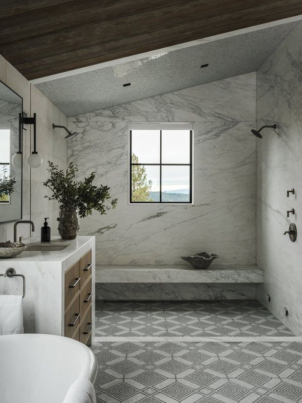 A refined contemporary bathroom featuring a marble-clad walk-in shower with a built-in bench, dual shower fixtures, a freestanding white bathtub, and a timber vanity with a pale stone top. Geometric patterned floor tiles contrast with the veined marble walls, while dark metal fittings and a large black-framed window add definition. A timber-lined ceiling and greenery provide warmth, with a partial scenic view visible through the window.