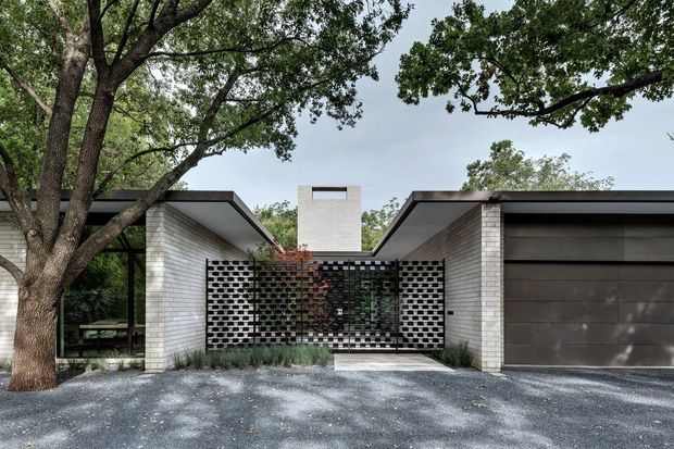 Single-storey modern residence viewed from the front, with low flat roof forms, pale brick walls, dark metal-framed glazing, a perforated brick screen at the entry courtyard, and an attached dark-panel garage. Mature trees frame the facade and a gravel driveway occupies the foreground.