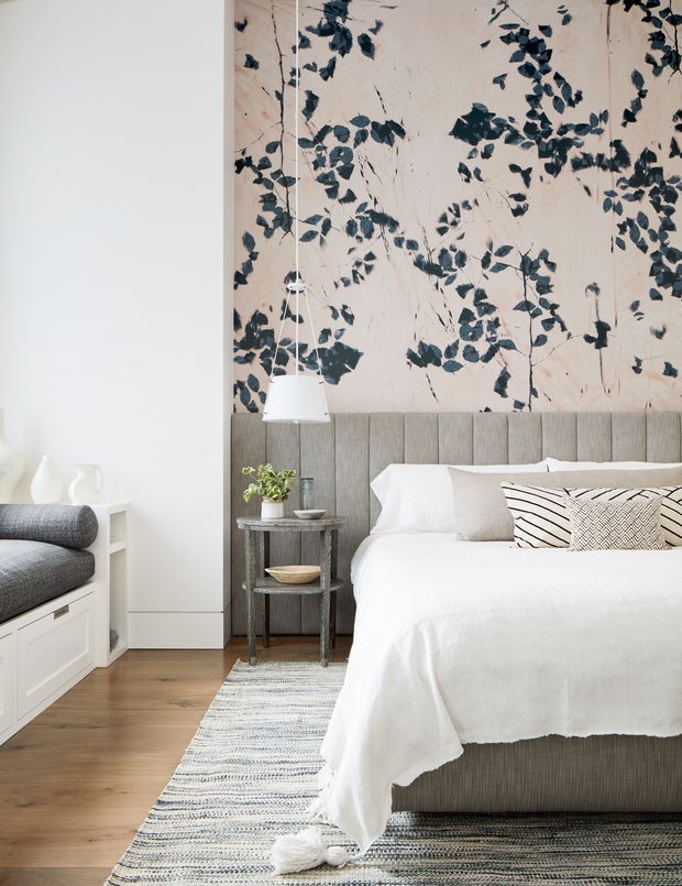 A softly styled contemporary bedroom with a large upholstered bed, white bedding, patterned cushions, and a dramatic botanical mural feature wall. The room includes a small round bedside table, pendant lights, timber flooring, a striped area rug, and built-in white cabinetry with a grey upholstered bench along the left wall.