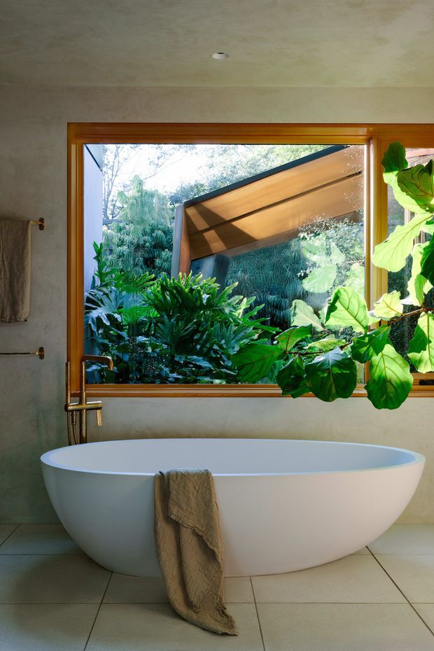 A serene contemporary bathroom featuring a freestanding oval bathtub positioned in front of a large timber-framed window. The window looks onto dense lush greenery and includes an angled external sunshade or awning. The room has smooth warm grey plaster walls, large pale floor tiles, a simple wall-mounted towel rail, and understated bath plumbing. A neutral textured towel hangs over the tub, while a large leafy plant occupies the right foreground.