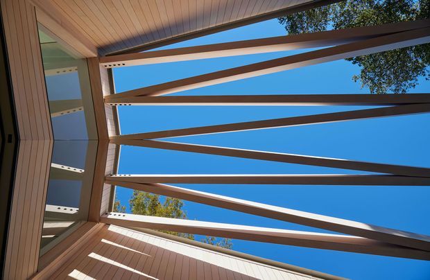 Upward-looking architectural view of a timber-lined covered outdoor space or courtyard, framed by angled and horizontal dark structural beams against a clear blue sky. Vertical timber wall cladding and a glazed opening are visible along the left side, with strong sunlight creating linear shadows across the ceiling and walls.
