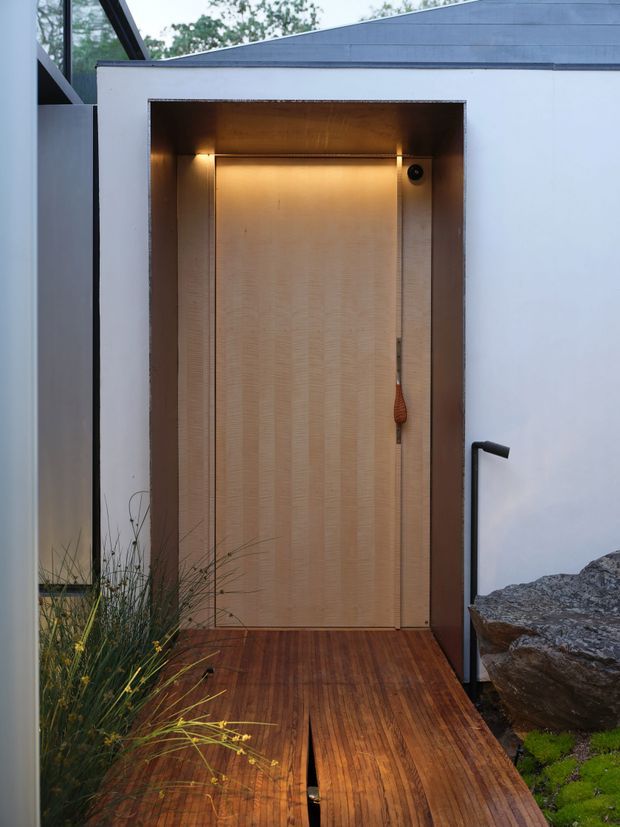 A contemporary exterior entry to a compact, box-like structure. A recessed timber-clad doorway is warmly illuminated from above and framed by dark metal or timber trim. The entry opens onto a narrow timber deck and walkway, with pale smooth exterior walls, angular roof geometry, exposed glazing at the left, and natural rocks and planting alongside.