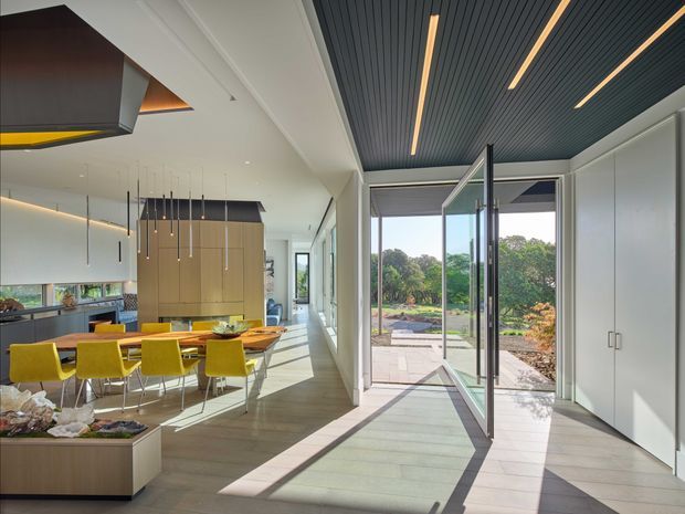 Bright contemporary open-plan interior combining a kitchen and dining area with a glazed connection to an outdoor garden terrace. The space features a large island with yellow dining chairs, pale timber cabinetry, integrated linear lighting, a dark slatted ceiling with recessed strip lights, extensive floor-to-ceiling glazing, and light timber or wood-look flooring. Sunlight creates strong geometric shadows across the floor, with landscaped grounds visible beyond the open glazed doors.