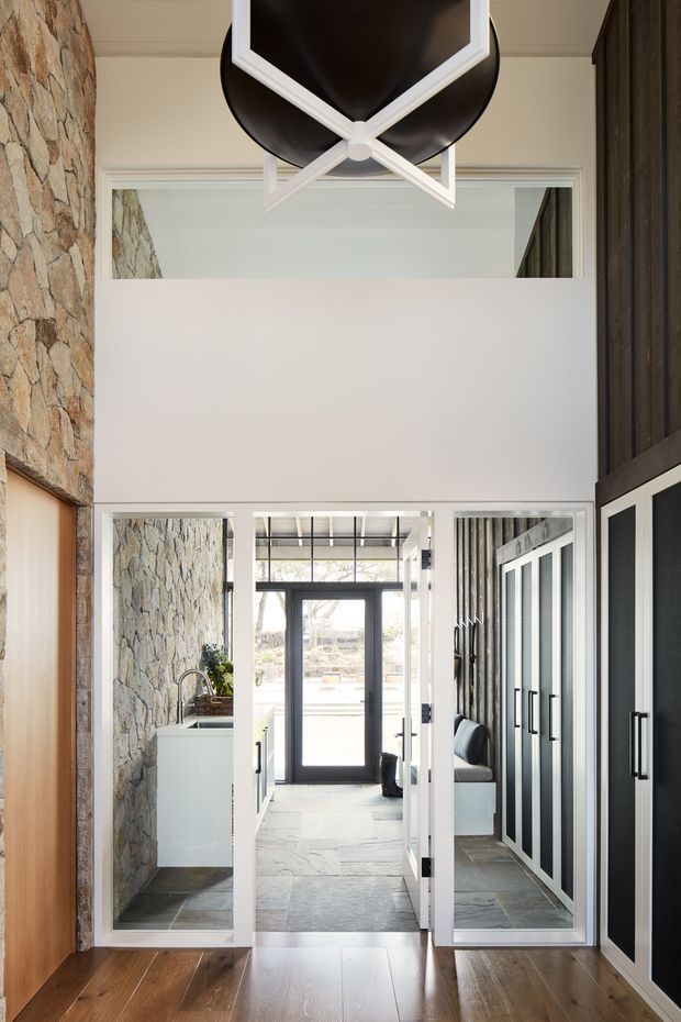 A bright, double-height residential entry hall with a dramatic oversized black-and-white geometric pendant light. The space combines warm timber flooring, a rough natural-stone feature wall, white-framed glazed internal doors, and dark built-in cabinetry with black handles. A small utility or kitchenette area and a seated nook are visible beyond the entry, with strong natural light entering through the far glazed door.