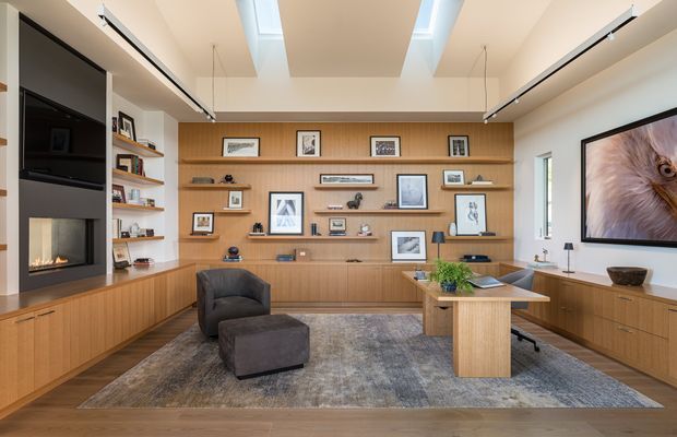 A spacious, refined living room with extensive mid-tone timber built-in cabinetry and open display shelving across the rear wall and lower side walls. The room features a large grey area rug, dark upholstered lounge chair and ottoman, a timber coffee table, concealed television cabinetry, a linear gas fireplace, artwork, and a compact desk or console area. Multiple roof skylights and clerestory-style openings provide abundant natural light, while slim black track lighting adds a contemporary architectural detail.