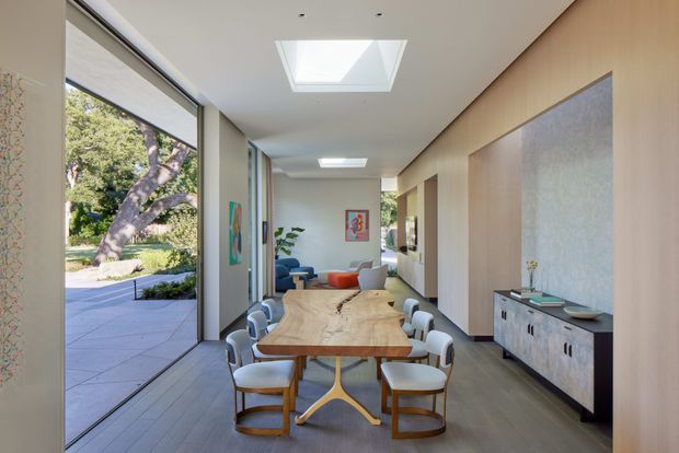 A bright, elongated open-plan dining and living area with a large timber dining table and upholstered dining chairs in the foreground. Full-height glazing opens toward a landscaped outdoor area, while a low timber sideboard and large textured wall panel line the right wall. Skylights bring additional daylight into the space, with a living area visible beyond the dining zone.