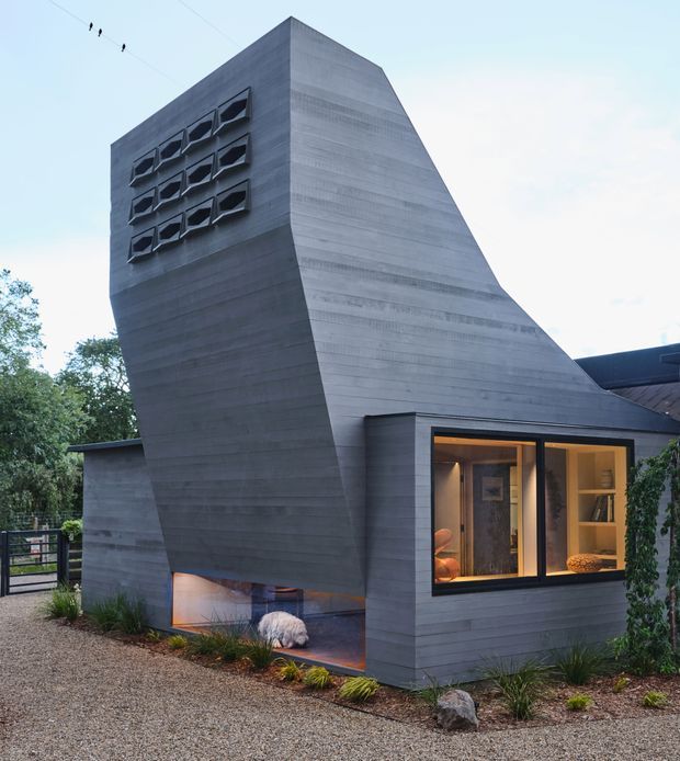 Exterior view of a sculptural contemporary house with a tall, sharply angled grey concrete volume, narrow geometric windows, and warm-lit floor-to-ceiling glazing at the lower level. The building sits in a landscaped gravel setting with planted borders and a gated walkway.