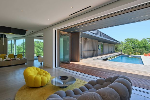 Contemporary open-plan living room opening directly onto a timber deck and rectangular swimming pool. The interior features floor-to-ceiling glazing, a sculptural yellow lounge chair, a rounded grey sofa, a low metallic coffee table, timber flooring, and a clean white ceiling with recessed linear air-conditioning vents. Outside, a separate pavilion-like structure has vertical timber cladding and a dark pitched roof.