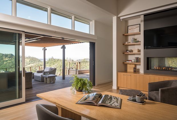 A bright contemporary living room with a large timber dining or work table in the foreground, built-in timber media cabinetry and open shelving, a wall-mounted television, and a linear gas fireplace. Full-height sliding glass doors open to a covered timber outdoor deck with seating and expansive green hillside views. High clerestory windows and the indoor-outdoor connection bring abundant natural light into the room.