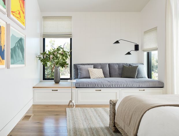 Bright, refined bedroom with a built-in window seat beneath two windows, featuring white paneled cabinetry, a grey upholstered cushion and pillows, pale timber flooring, a soft neutral rug, and a partially visible neatly made bed. Abstract framed artwork and a sculptural wall-mounted black reading light add restrained decorative accents.