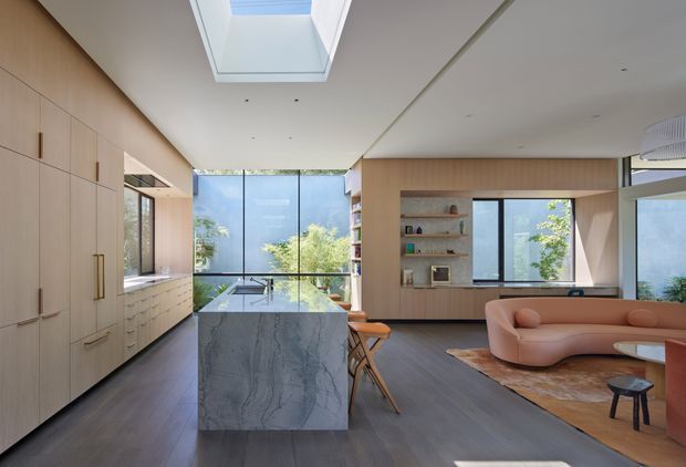 Open-plan contemporary kitchen and living area with extensive light timber cabinetry, a large veined stone island, dark timber flooring, built-in shelving, and a curved blush sofa. Floor-to-ceiling glazing and a large skylight bring in abundant natural light, with greenery visible outside.