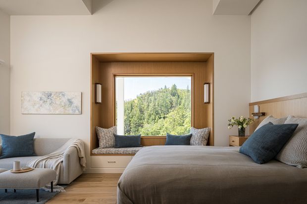 A calm, softly lit bedroom with a large upholstered bed in the foreground and a built-in timber-framed window seat overlooking a wooded landscape. The room features warm timber flooring, a neutral upholstered sofa, layered grey and blue cushions, pale walls, integrated bedside storage, and simple wall-mounted lighting. A shallow ceiling bulkhead and recessed window surround add architectural definition.