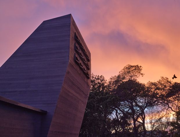 A dramatic exterior architectural photograph at sunset, showing a tall sculptural building volume with sharply angled, monolithic walls and a narrow vertical strip of recessed windows. The facade appears dark grey to muted brown with horizontal surface banding, silhouetted against a vivid pink, purple, and orange sky, with mature trees framing the right side.