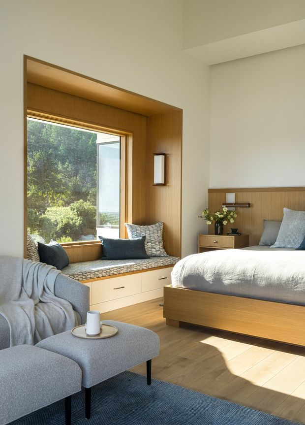 A warm, light-filled bedroom with a built-in timber window seat and integrated drawers, a timber bed with upholstered bedding, a matching bedside cabinet, and a grey upholstered lounge chair and ottoman. The room features pale walls, timber flooring, blue-grey textiles, and a large window overlooking greenery. Built-in timber panelling frames the window and continues behind the bed, with a simple wall sconce providing understated lighting.