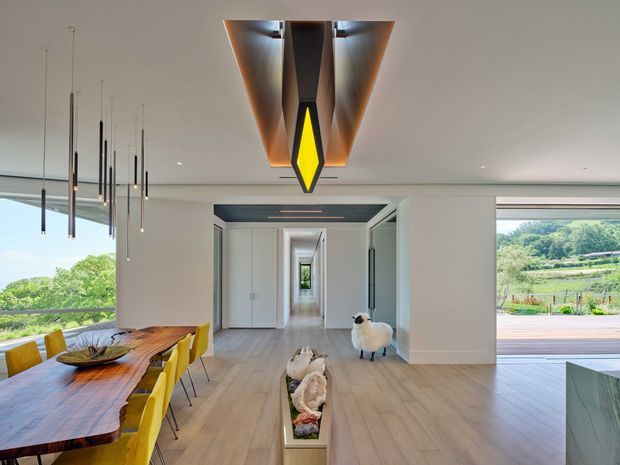 Bright, open-plan contemporary dining and living space with pale timber flooring, white walls, expansive glazing, and views toward a green rural landscape. A sculptural live-edge timber dining table with yellow chairs occupies the left side, while a dramatic geometric timber-and-metal ceiling light forms a central feature. The room opens through a long, carefully framed hallway toward a distant window, with a partial kitchen island visible at right and a deck beyond the glazing.