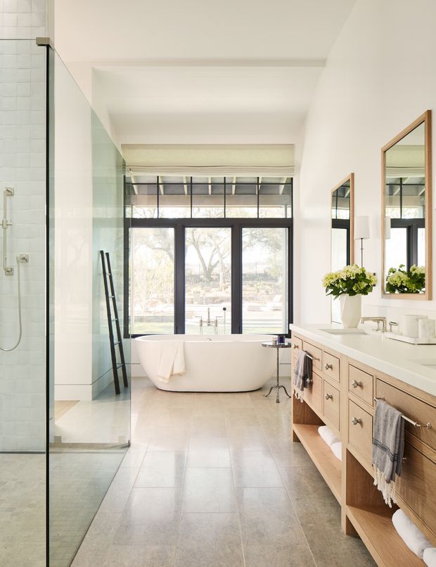 Bright, spacious bathroom with a freestanding white bathtub positioned in front of large black-framed windows, a glass-enclosed walk-in shower on the left, and a long light-wood double vanity with white countertop and mirrors on the right. The room has pale tiled flooring, white walls and ceiling, abundant natural light, and a clean refined transitional feel.