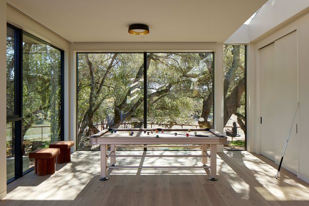 A bright, glass-walled recreational room surrounded by mature trees, featuring a central foosball table, two small upholstered stools, pale timber flooring, built-in light-coloured cabinetry, and a skylight or clerestory opening. Large black-framed windows provide strong views into the landscaped setting.
