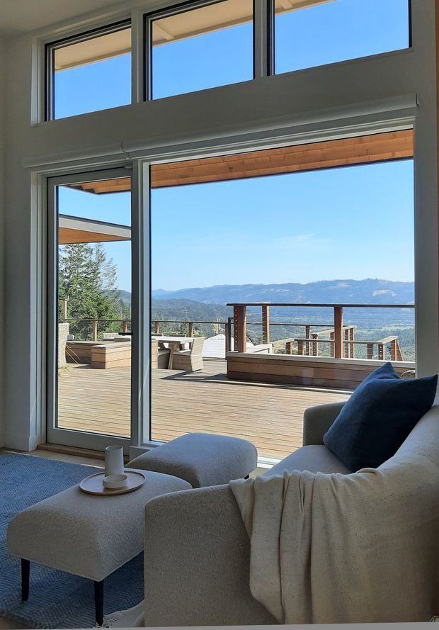 A bright contemporary living room opens through extensive floor-to-ceiling glazing onto a large timber deck and outdoor terrace. The room contains a pale upholstered sofa and ottoman, a dark blue cushion, a light throw, and a small round tray table. The glazing frames expansive mountain and valley views, while the deck has timber flooring, timber-and-metal balustrades, and outdoor seating. High-level windows and a covered roof edge create a light-filled, elevated setting.