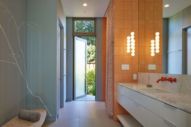 A refined contemporary bathroom viewed toward an open exterior door. The room features a long floating white vanity with integrated drawers, a pale marble-look benchtop and backsplash, twin basins with red wall-mounted taps, and vertical round-bulb sconces set against a warm orange tiled feature wall. Cool blue-grey vertically grooved wall panelling, a patterned botanical wall surface, large-format floor tiles, and a small upholstered bench with a rolled towel complete the space. Natural greenery is visible beyond the doorway.