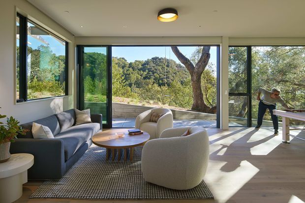 Bright contemporary living room with expansive floor-to-ceiling glazing framing a leafy landscape. The room features a low grey sofa, sculptural cream lounge chairs, a round timber coffee table, a large patterned rug, pale timber flooring, white walls and a dark-framed glazed door opening to an outdoor terrace. Strong sunlight creates long shadows across the floor, with a person visible at a table near the right-hand glazing.