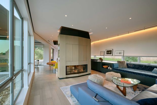 A spacious contemporary living room with full-height glazing along one side, a sculptural central fireplace clad in pale cabinetry, and a large blue sectional sofa arranged around a low oval glass coffee table. Warm concealed and recessed lighting, pale timber flooring, dark built-in joinery, and framed artwork create a refined, open-plan interior. A dining or kitchen area is visible beyond the fireplace.