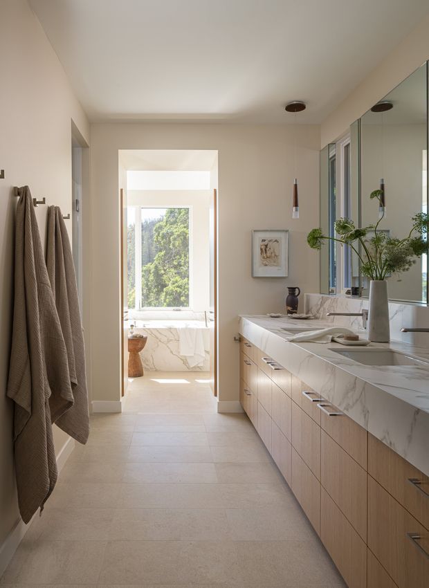 A bright, elongated contemporary bathroom with a double vanity along the right wall, featuring pale timber cabinetry, a veined white stone benchtop and integrated backsplash, large mirrored wall panels, and twin basins. Neutral beige walls and large-format light floor tiles create a calm, refined atmosphere. Towels hang from wall hooks on the left, while an open doorway leads to a separate bathing area with a freestanding bathtub, marble-look surfaces, and a large window overlooking greenery.