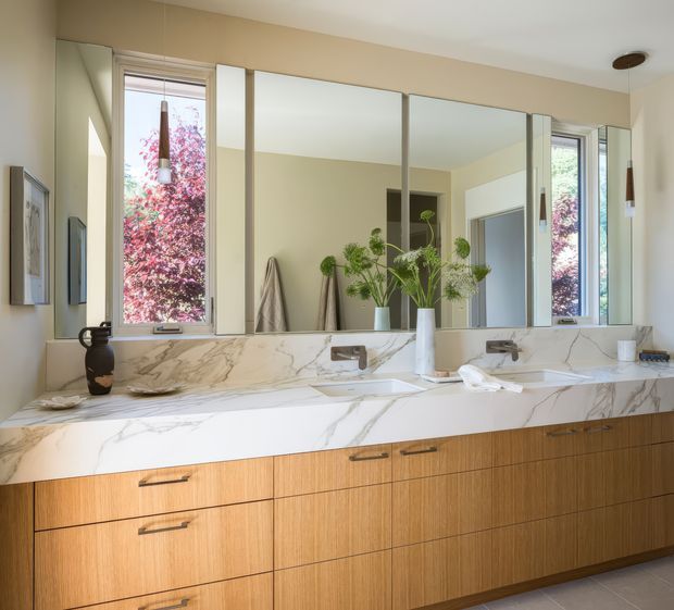 Bright, refined bathroom vanity with a long timber drawer cabinet, white marble-look stone benchtop and integrated backsplash, twin wall-mounted basins, large frameless mirror panels, and tall windows. Warm neutral walls and natural light create an elegant, calm interior.