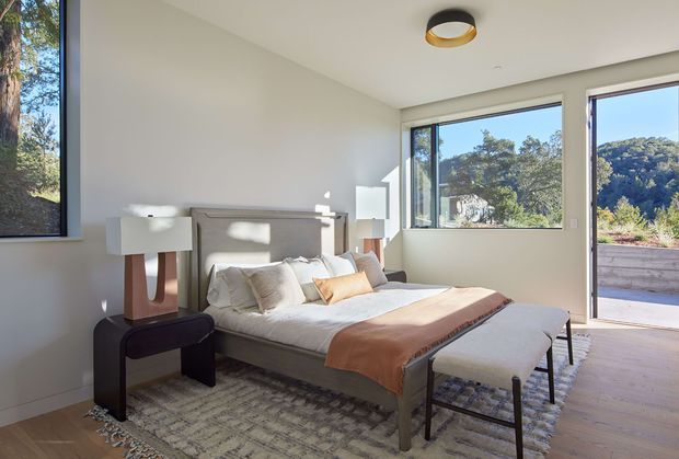 A bright, contemporary bedroom with a low upholstered bed, layered neutral bedding, a warm brown throw, and a pair of bedside tables with lamps. Large windows and a glazed door provide abundant natural light and views toward a wooded landscape. The room has pale walls, light timber flooring, a textured neutral rug, and a streamlined circular ceiling light.