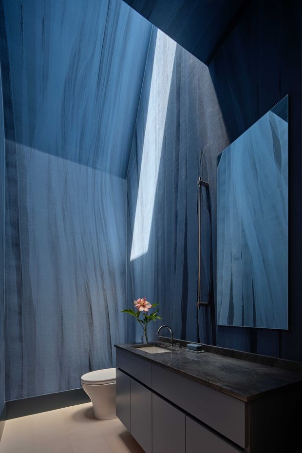 A dramatic, compact bathroom with deep blue vertically textured wall finishes, a dark floating vanity with integrated basin and dark stone benchtop, wall-mounted mirror, sculptural pendant or wall light, and a white toilet. A narrow skylight or high opening casts a strong beam of natural light into the space, creating a moody, luxurious atmosphere.