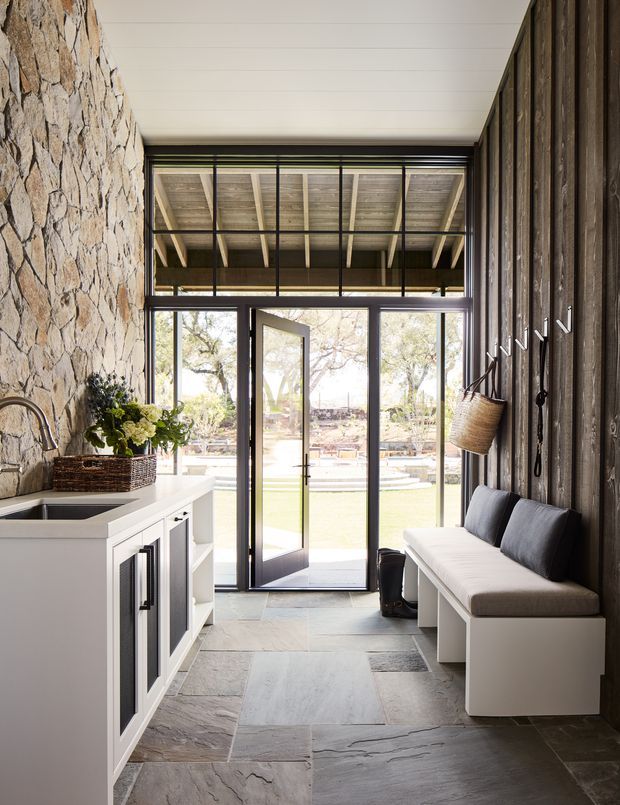 A bright, compact entryway with a built-in mudroom and utility joinery. The space features a rough natural-stone wall, dark vertical timber wall panelling, large black-framed glazed doors opening to a lawn, a white built-in cabinet with sink, and a white bench seat with grey cushions. Slate-like stone flooring and an exposed timber-lined ceiling reinforce the rustic, natural character.