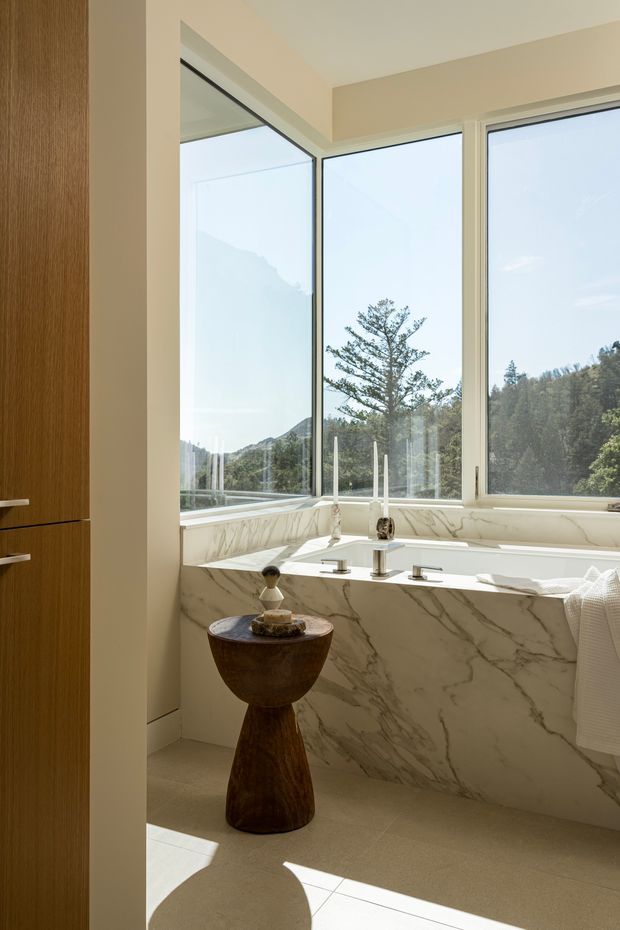 Bright, refined bathroom featuring a freestanding bathtub enclosed by a dramatic veined marble surround and ledge, with large corner windows overlooking a wooded landscape. A warm timber vanity or storage unit is visible at left, while a sculptural dark wood pedestal table and understated bath accessories add contrast. Natural light creates a calm, luxurious atmosphere.