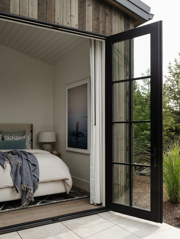 A refined bedroom opening directly onto a landscaped garden through a fully open black-framed glazed door. The room has soft neutral walls, a white paneled ceiling, light timber flooring, a neatly styled upholstered bed with layered bedding, curtains, bedside table and wall artwork. The exterior facade is clad in vertical weathered timber boards, while the threshold and terrace are finished in large pale tiles.