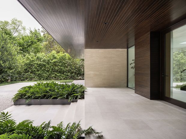 A refined contemporary covered entry terrace beside a landscaped garden. Large pale stone or porcelain floor tiles continue across the open-air space, with a dark timber-lined soffit and matching timber-clad wall framing a large glazed doorway. A pale textured masonry wall, built-in planter beds, and lush ferns create a calm indoor-outdoor transition.