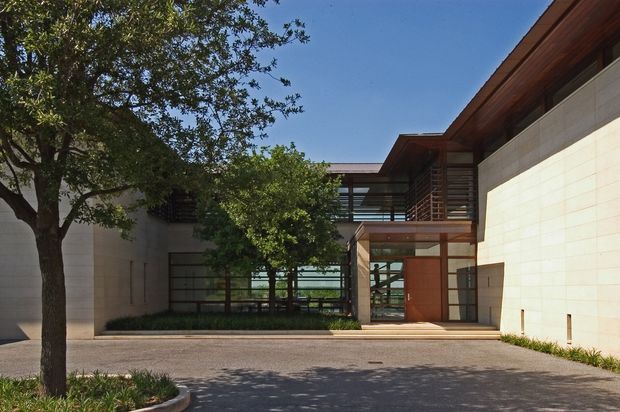 A contemporary multi-level building facade arranged around a landscaped courtyard or entry drive. The composition features pale masonry walls, extensive dark-framed glazing, warm timber roof soffits and balcony screens, and a recessed glazed entrance beneath a projecting upper level. Mature trees and low planting soften the paved forecourt.