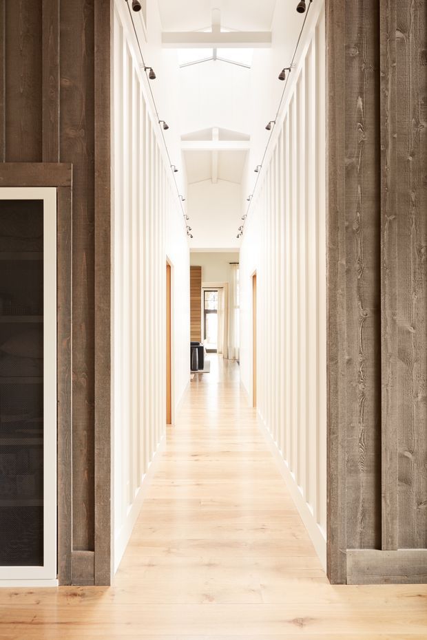 A bright, narrow residential hallway viewed through a wide opening framed by vertical reclaimed-looking timber boards. The corridor has pale timber flooring, white vertically fluted or slatted wall detailing, multiple timber door openings, and a high white pitched ceiling with exposed beams and linear track lighting. A dark built-in display or storage cabinet is partially visible at the left edge. The space feels airy, warm, and contemporary with strong axial perspective.
