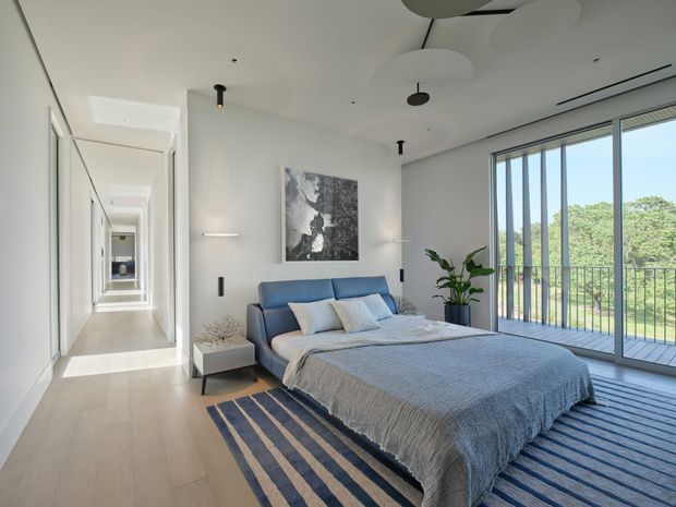 Bright contemporary bedroom with a low-profile blue upholstered bed, layered neutral bedding, striped rug, minimalist bedside table, indoor plant, and abstract wall art. Full-height sliding glazing opens to a balcony or terrace with vertical privacy screens and leafy views. A long hallway and additional rooms extend beyond the bedroom, while sculptural circular ceiling fixtures and recessed lighting reinforce the clean modern design.