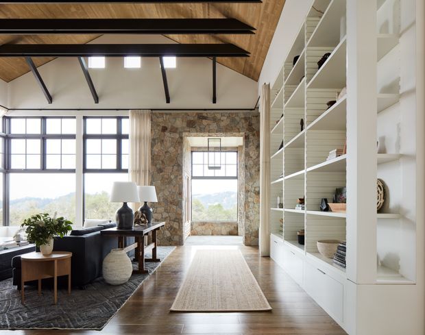 Bright, refined living room with a high pitched timber-lined ceiling, exposed dark beams, expansive black-framed windows, a substantial stone-clad feature wall, and full-height white built-in shelving. The room includes a dark sofa, console, lamps, rugs, and a large central window with scenic views beyond.