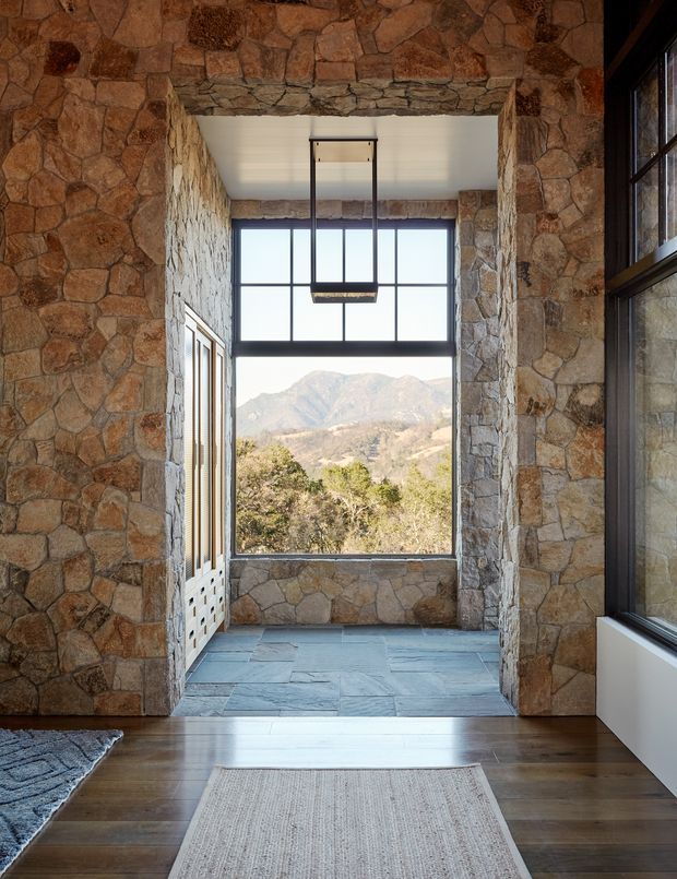 A rustic mountain-view interior passage or entry space framed by extensive irregular natural-stone walls and a heavy stone lintel. Dark-framed clerestory-style glazing and a large window overlook a rugged mountain landscape, while a simple suspended rectangular light hangs above a slate-finished floor. The foreground has dark timber flooring and a neutral woven rug, with additional dark-framed glazing on the right.