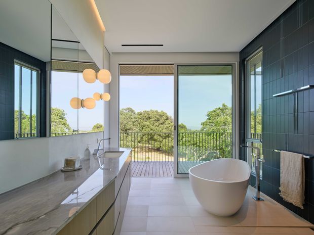 Bright contemporary bathroom with a freestanding oval bathtub, floating vanity, large stone benchtop, wall-mounted basin tapware, and dark vertically tiled feature walls. Full-height glazing and a glass door open to a timber balcony overlooking leafy surroundings. Round pendant lights and a large mirror add visual interest.