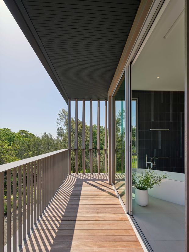 A contemporary upper-level balcony or terrace with warm timber decking, slender vertical balustrade and privacy screens, and a deep dark-lined roof overhang. Full-height glazed doors open into a modern bathroom or interior space with dark tiled walls and a freestanding basin visible inside. Strong linear shadows from the balustrade fall across the deck, with leafy greenery beyond.