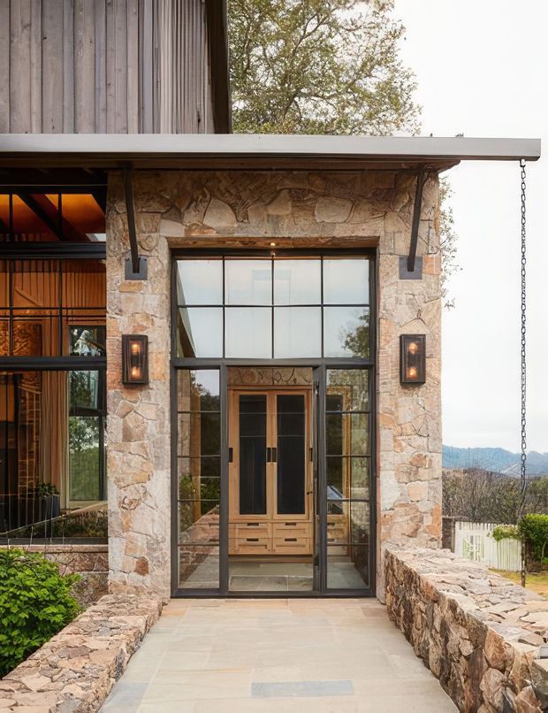 Exterior view of a refined stone-clad entry facade with a covered porch and broad flat roof overhang. A framed glazed entry door is set within a black metal window wall, with timber cabinetry visible inside. Rough natural stone walls, timber vertical cladding, exposed dark metal supports, wall-mounted lanterns, and a suspended rain chain define the entrance. A paved walkway is bordered by low stone walls, with trees and distant hills visible beyond.