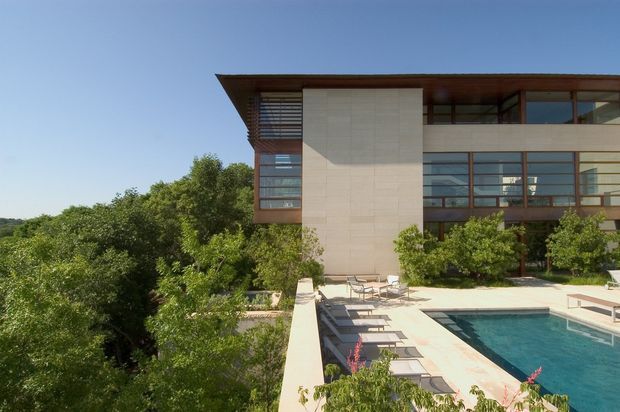 Exterior view of a contemporary multi-storey residence overlooking a landscaped pool terrace. The building features broad horizontal glazing with dark timber or brown-toned frames, a projecting flat roof, a large pale stone-clad vertical wall, and horizontal external sun-shading elements. A rectangular swimming pool, paved deck, loungers, planted borders, and mature trees occupy the foreground.