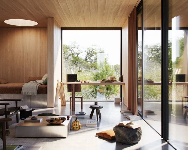 A refined open-plan interior with a compact living area, integrated sleeping nook, and a simple work or dining table positioned beside full-height glazing. Warm timber wall and ceiling panelling, dark slender window frames, pale flooring, and sculptural low furniture create a calm contemporary atmosphere. Garden vegetation is visible through the glazing.