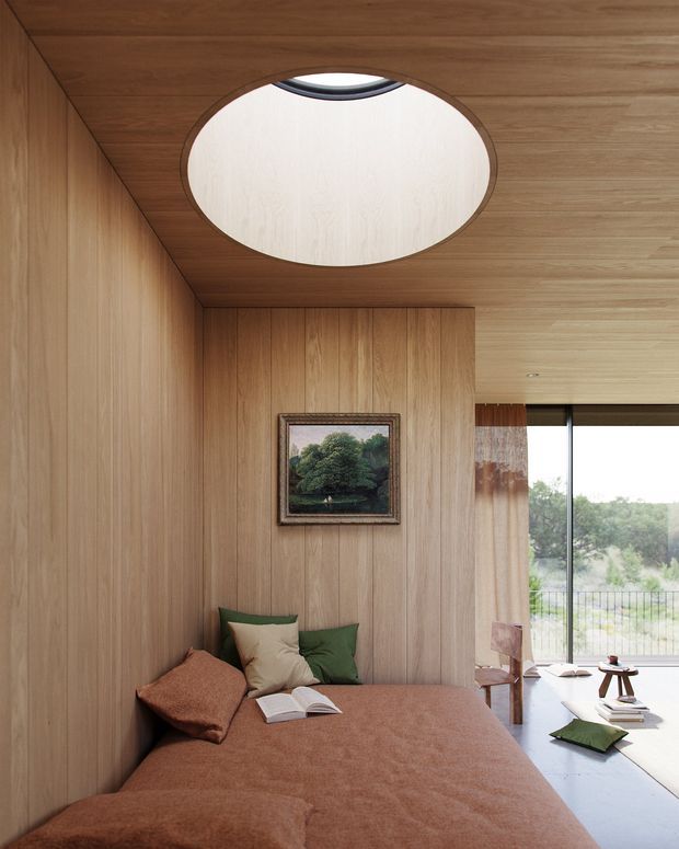 A compact, highly enveloped bedroom or sleeping nook clad in warm vertical timber panelling across the walls and ceiling. A large oval roof skylight brings in daylight, while a full-height glazed opening at right overlooks greenery. The room contains a low bed with rust-brown bedding and cushions, a framed landscape artwork, and a small seating or reading area near the window. The design combines rich timber surfaces, soft earthy textiles, and strong natural light.