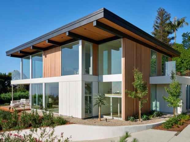 A contemporary two-storey residence with a dramatic flat roof and deep projecting eaves. The facade combines vertical warm timber cladding, white panelled walls, dark roof fascia, extensive floor-to-ceiling glazing, and projecting upper-level balconies. It is set within a landscaped garden with paved paths and planting under clear blue skies.