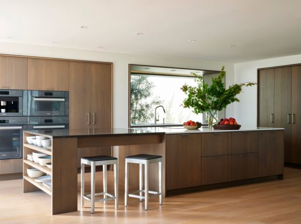 Contemporary kitchen with extensive dark timber cabinetry, integrated black appliances, a large central island with a raised breakfast bar, and two stools. A wide window above the sink brings in natural light, while open shelving displays white crockery and fruit bowls and a tall interior plant adds greenery. The room has a warm, refined, clean appearance with pale timber flooring and a simple white ceiling.