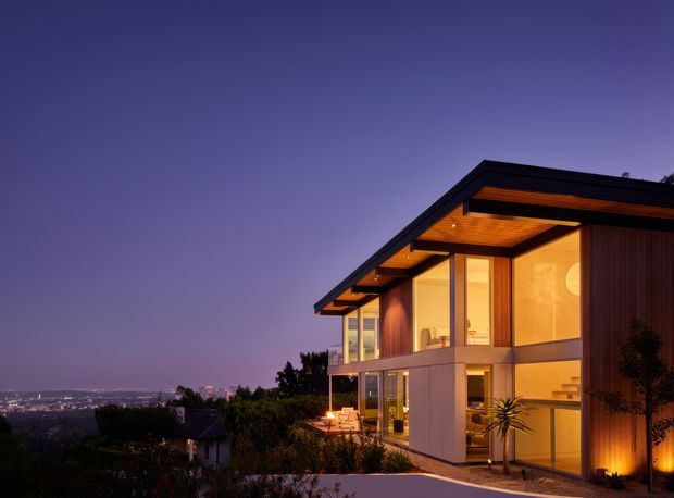 Twilight exterior view of a contemporary multi-level residence on a sloping site, with expansive floor-to-ceiling glazing, warm illuminated interiors, dark roof planes, timber soffit detailing, and vertical timber cladding. The house overlooks a distant urban landscape and incorporates a projecting upper-level form with sheltered outdoor areas.