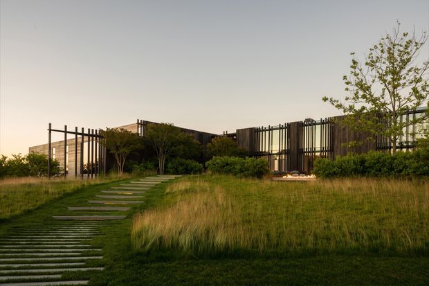 Single-storey contemporary house set within a landscaped grassland garden. The low, horizontally arranged building has extensive glazing and dark vertical timber or metal screening elements forming open, rhythmically spaced facades and courtyards. A stepping-stone path leads through tall ornamental grasses toward the entrance, with mature planting partially obscuring the building. The scene is photographed in warm, subdued evening light.