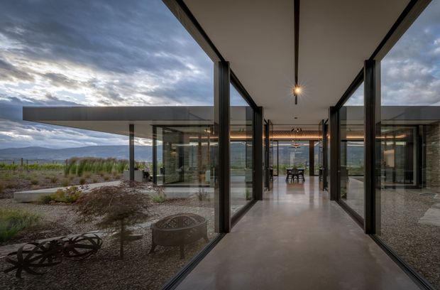 A dramatic contemporary single-level home viewed from a covered glazed passageway. Floor-to-ceiling glass walls and slim dark metal frames line both sides, opening to a minimalist gravel landscape and distant mountain scenery. The central corridor leads toward a dining area, with exposed dark structural beams, a smooth pale concrete floor, and a restrained linear pendant light. The architecture emphasizes transparency, long horizontal roof forms, and indoor-outdoor connection.